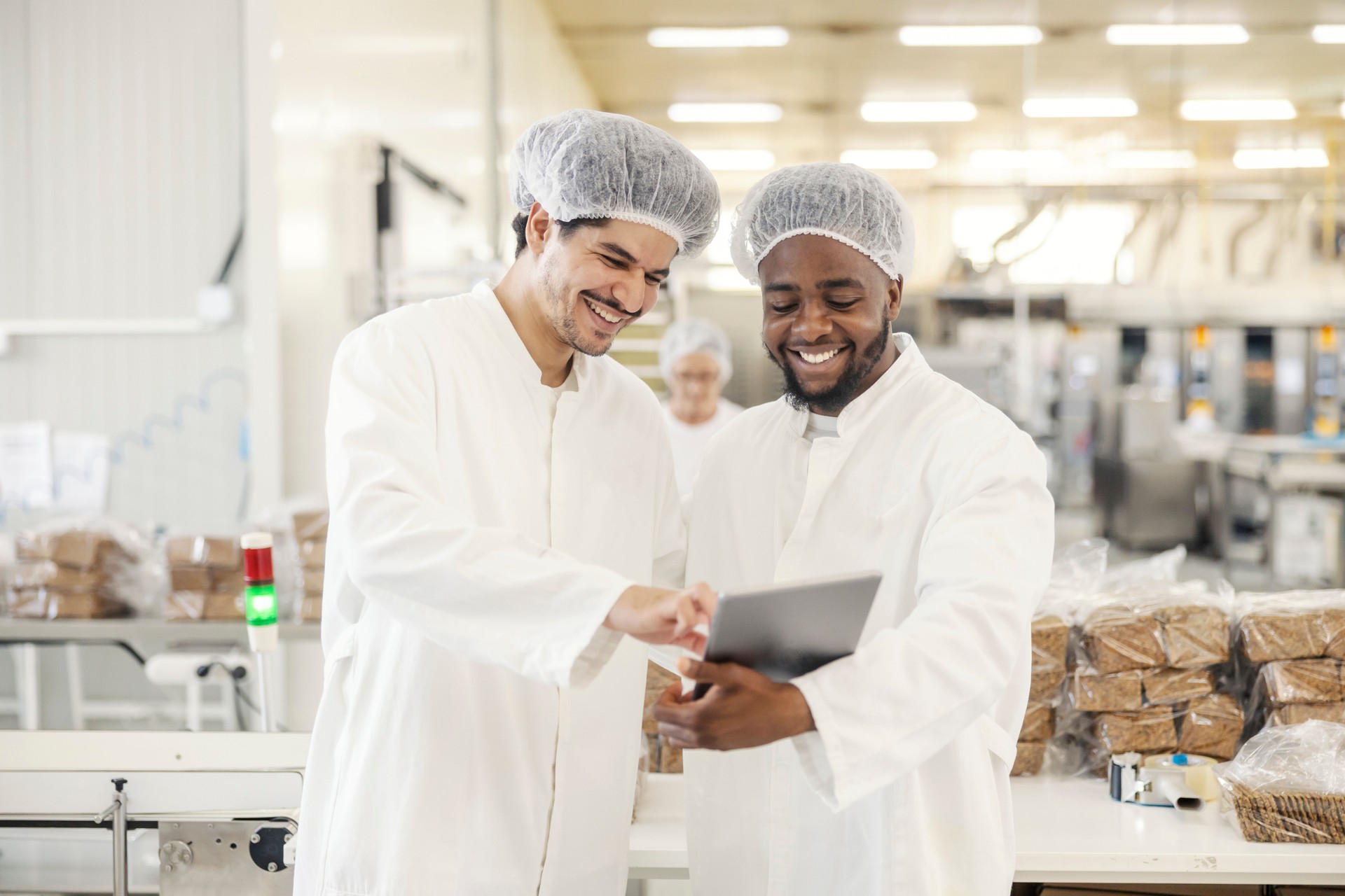 Two interracial supervisors scrolling and smiling at tablet at food factory.